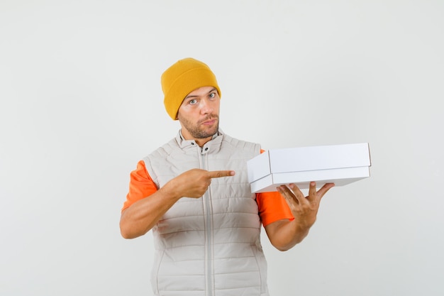 Young man pointing at cardboard box in t-shirt, jacket, hat front view.