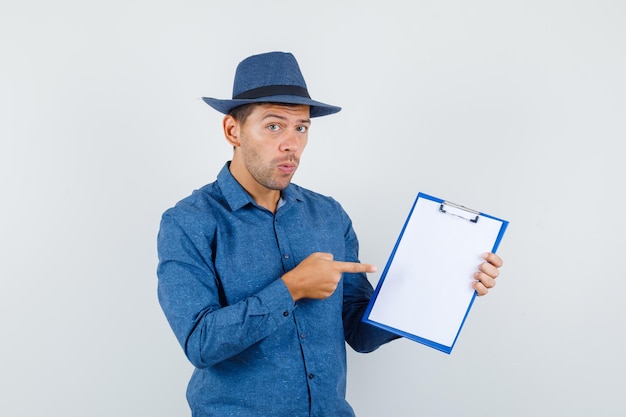 Young man pointing at clipboard in blue shirt, hat and looking careful , front view.