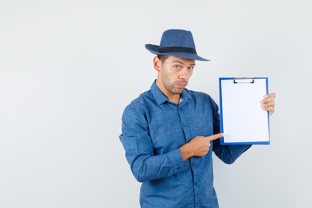 Young man pointing at clipboard in blue shirt, hat and looking serious , front view.