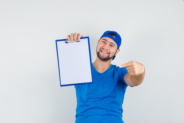 Young man pointing at clipboard in blue t-shirt and cap and looking merry