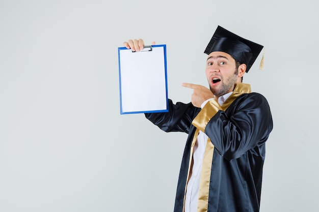 Young man pointing at clipboard in graduate uniform and looking happy. front view.