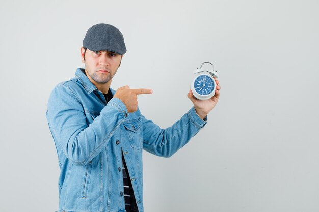 Young man pointing at clock in jacket,cap and looking serious. 