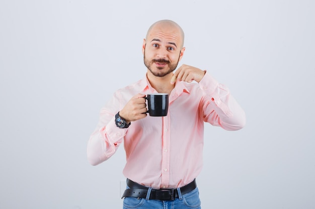 Young man pointing at cup in pink shirt,jeans and looking alarmed , front view.