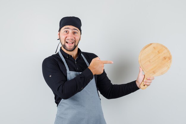 Young man pointing at cutting board in shirt, apron and looking cheerful , front view.