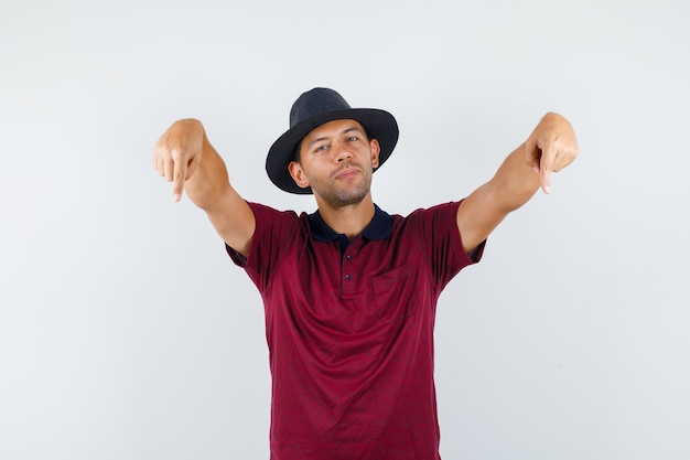 Free photo young man pointing down in t-shirt, hat and looking confident , front view.