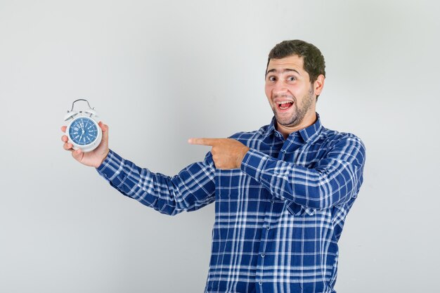 young man pointing finger at alarm clock in checked shirt and looking excited.