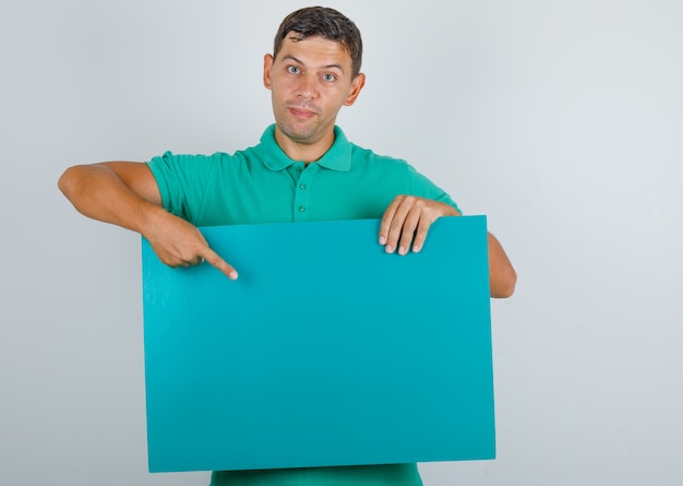 Молодой человек указывая пальцем на синий плакат в зеленой футболке, вид спереди.