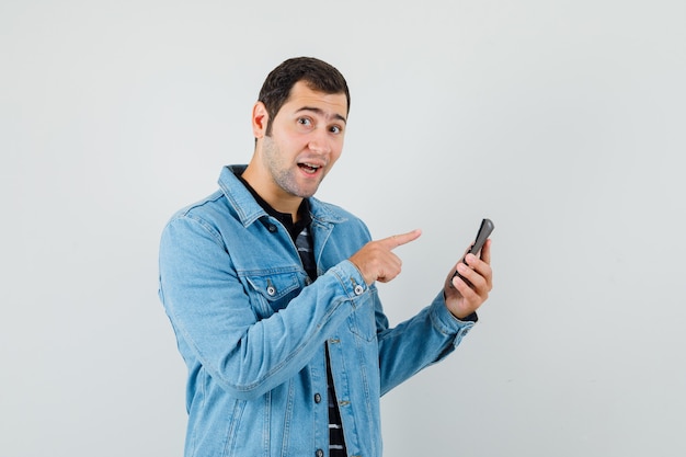 Young man pointing finger at calculator in t-shirt, jacket and looking cheerful , front view.