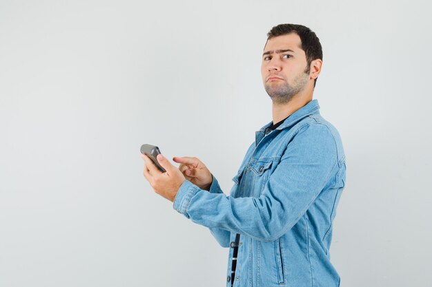 Young man pointing finger at calculator in t-shirt, jacket and looking serious. .