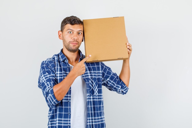 Young man pointing finger at cardboard box in shirt and looking excited , front view.