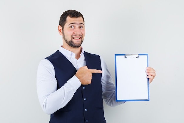 Young man pointing finger at clipboard in shirt, vest and looking cheery