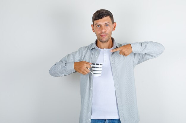 Young man pointing finger at cup of drink in shirt, jeans and looking glad