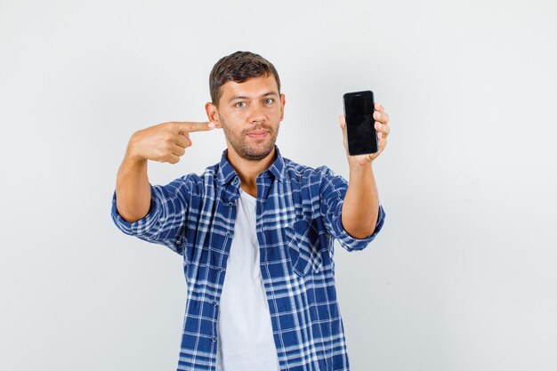 Молодой человек указывая пальцем на смартфон и улыбается в вид спереди рубашку.