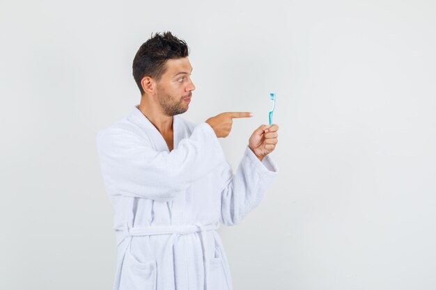 Young man pointing finger at toothbrush in white bathrobe , front view.