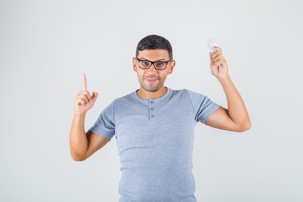 Young man pointing finger up and holding light bulb in grey t-shirt, glasses front view.