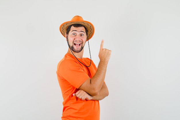 Young man pointing finger up in orange t-shirt, hat and looking merry. front view.