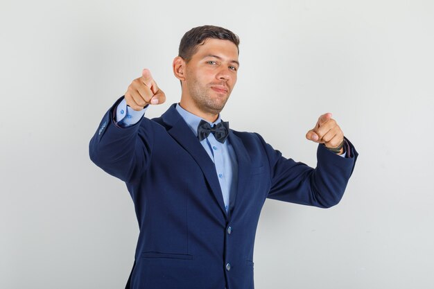 Young man pointing fingers to camera in suit and looking cheerful.