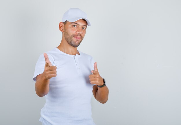 Young man pointing fingers at camera in white t-shirt, cap and looking confident, front view.