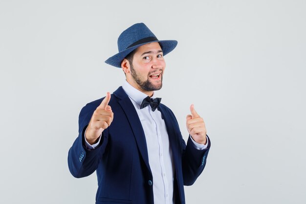 Young man pointing fingers at front in suit, hat and looking cheerful , front view.