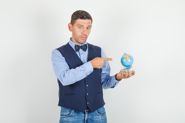 Foto gratuita giovane uomo che punta le dita al globo in tuta, jeans e guardando attento.