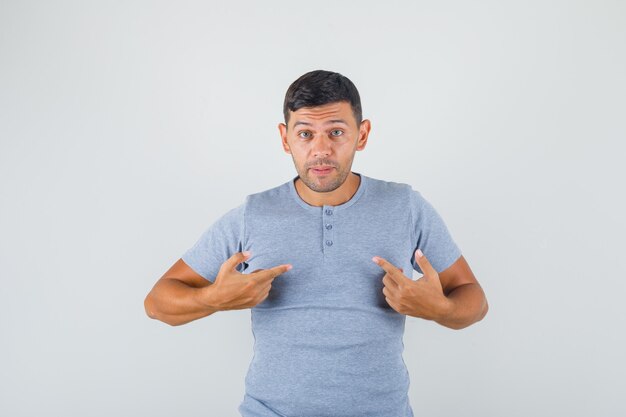 Young man pointing fingers at himself and asking 'me?' in grey t-shirt and looking doubted