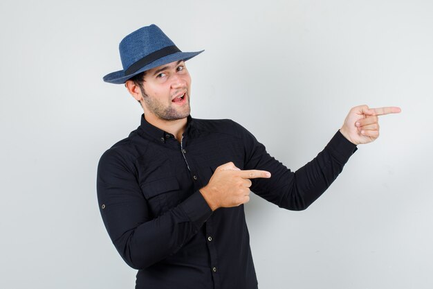 Young man pointing fingers to side in black shirt, hat and looking merry.