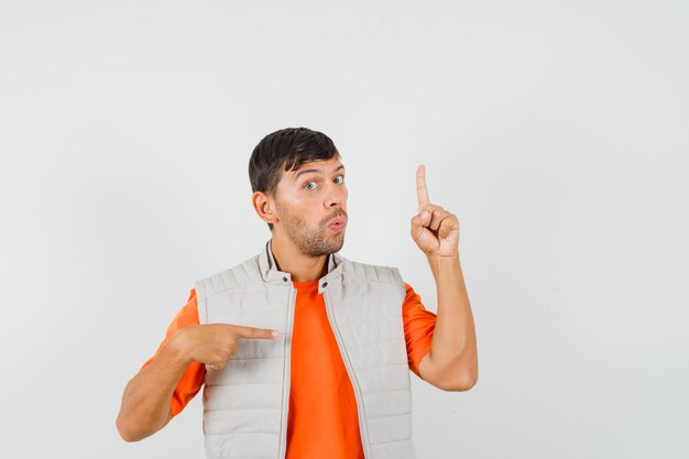 Young man pointing fingers up and aside in t-shirt, jacket and looking curious. front view.