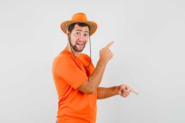 Молодой человек указывая пальцами вверх и вниз в оранжевой футболке, шляпе и радостно глядя, вид спереди.