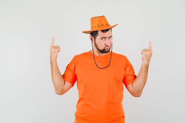 Giovane uomo che punta le dita in alto in maglietta arancione, cappello e guardando dubbioso, vista frontale.