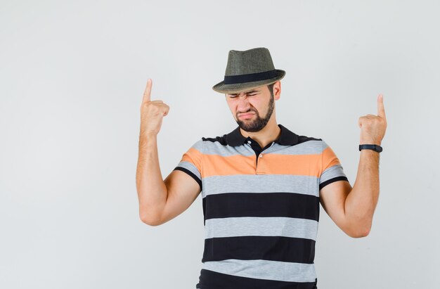 Young man pointing fingers up in t-shirt, hat and looking hopeless , front view.