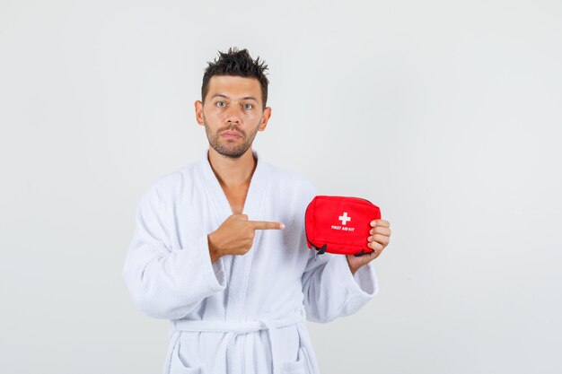 Young man pointing at first aid kit in white bathrobe and looking serious. front view.