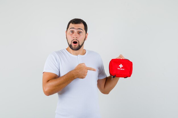 Young man pointing at first aid kit in white t-shirt and looking alarmed , front view.