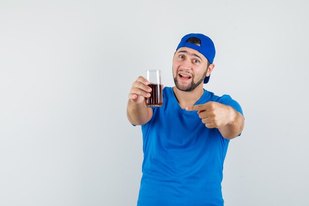 青いTシャツとキャップの飲み物のガラスを指して楽観的に見える若い男
