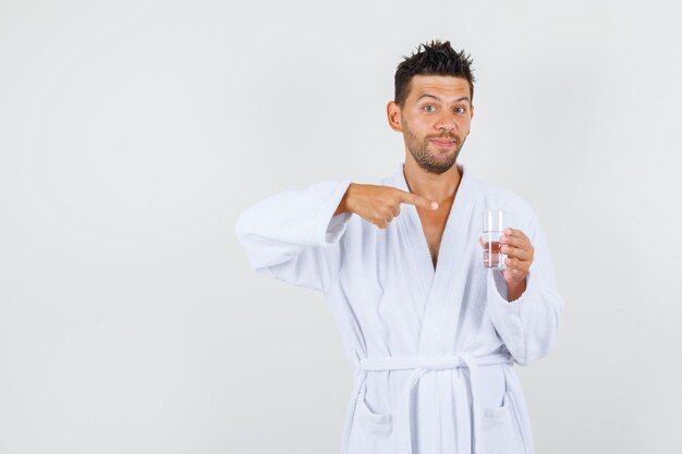 Young man pointing at glass of water in white bathrobe and looking joyful. front view.