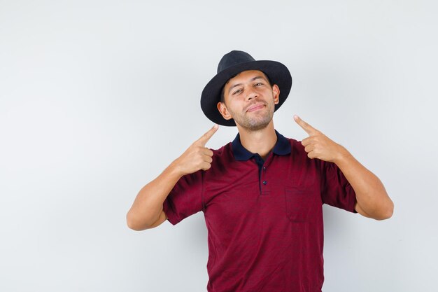 Young man pointing at himself in t-shirt, hat and looking cheerful. front view.