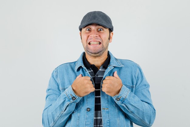 Free photo young man pointing at himself with clenched teeth in t-shirt, jacket, cap and looking surprised