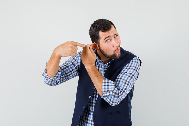 Young man pointing at his ear pulled by finger in shirt, vest , front view.