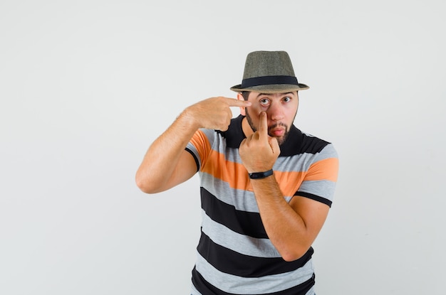 Young man pointing at his eyelid pulled by finger in t-shirt, hat , front view.