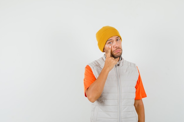 Young man pointing at his lower eyelid in t-shirt, jacket, hat and looking sad. front view.