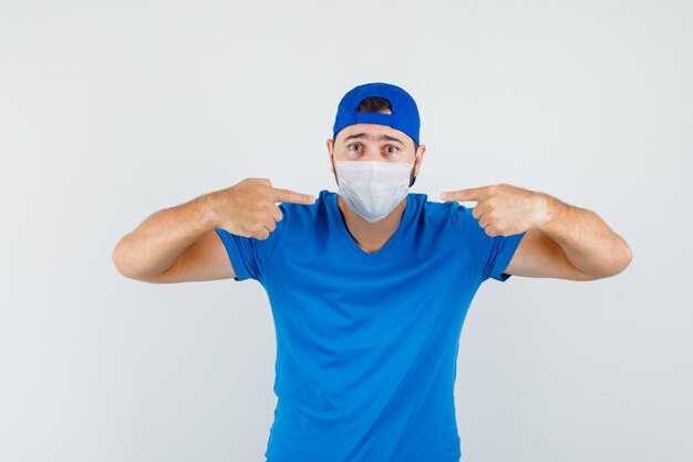 青いTシャツとキャップで彼の医療マスクを指して注意深く見ている若い男