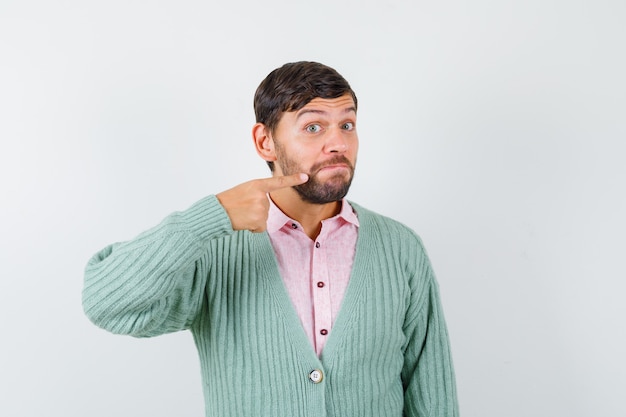 Young man pointing at his mouth in shirt, cardigan and looking puzzled. front view.