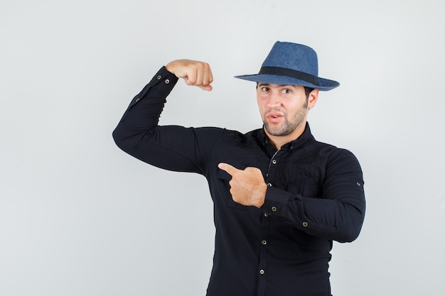 Giovane che punta al suo muscolo in camicia nera, cappello e sembra forte.