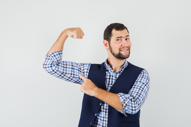 Young man pointing at his muscle in shirt, vest and looking confident , front view.