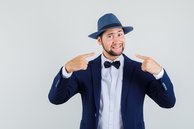 Giovane che indica al suo sorriso in vestito, cappello e che sembra allegro, vista frontale.