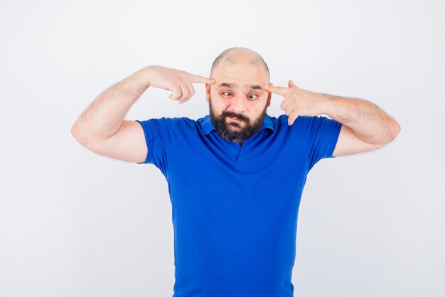 Young man pointing at his squinted eyes in blue shirt and looking weird. front view.