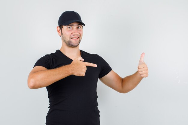 Young man pointing at his thumb up in black t-shirt, cap and looking cheery