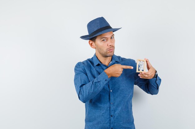 Young man pointing at hourglass in blue shirt, hat and looking sensible , front view.