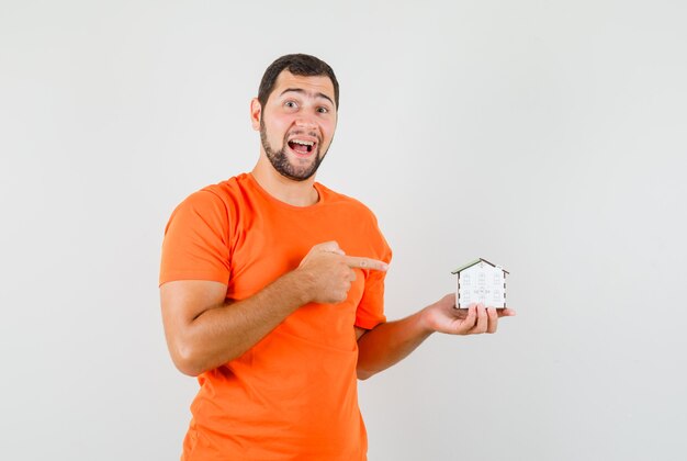 Young man pointing at house model in orange t-shirt and looking merry. front view.