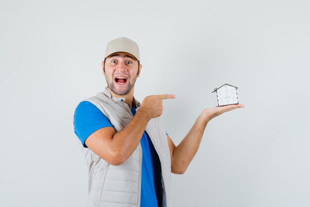 Young man pointing at house model in t-shirt, jacket, cap and looking happy , front view.