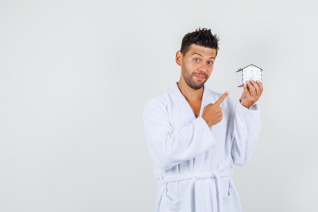 Young man pointing at house model in white bathrobe and looking curious , front view.